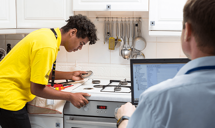 Young gas engineer leaning over a gas hob with an inspector watching him from behind a laptop